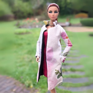 May include: A fashion doll wearing a white coat with pink and green floral patterns, a red dress, and a silver necklace. The doll has red hair and brown eyes.