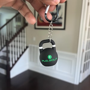 May include: A black kettlebell keychain with white stripes and the text "R.G. GOLF" in green. The keychain is attached to a black carabiner clip with a silver accent.