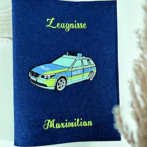Könnte beinhalten: Ein marineblauer Filz-Dokumentenhalter mit den gestickten Wörtern "Zeugnisse" und "Maximilian" in grüner Schrift. Ein farbenfrohes gesticktes Polizeiauto ist mittig auf der Vorderseite.