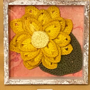 May include: A framed crocheted artwork featuring a yellow flower with a smaller yellow center and a green crocheted leaf. The artwork is displayed on a pink background.