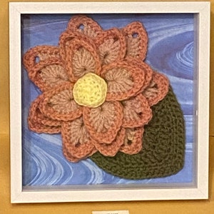 May include: A framed crocheted flower with pink and yellow petals and a green leaf. The flower is displayed on a blue background with a wavy pattern.