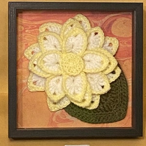 May include: A framed crocheted flower with yellow and white petals and a green crocheted leaf. The flower is displayed on a marbled background.
