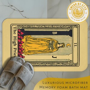 May include: A beige bath mat with a tarot card design featuring a woman in a yellow robe holding a book. The card is titled "The High Priestess" and has the number "II" in the corner. The mat has the text "LUXURIOUS MICROFIBER MEMORY FOAM BATH MAT" at the bottom.