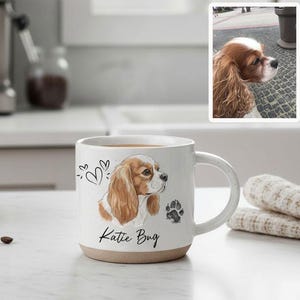 May include: White ceramic mug with a brown base, featuring a Cavalier King Charles Spaniel illustration, hearts, and a paw print. The mug has the name "Katie Bug" printed on it. Coffee beans and a coffee maker are in the background.