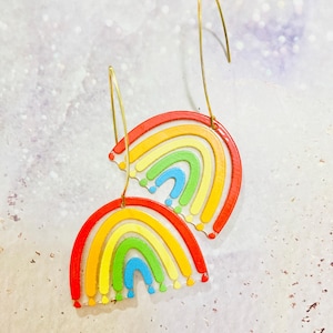 Peut inclure: Une paire de boucles d'oreilles pendantes avec un motif arc-en-ciel en demi-cercle. Chaque boucle présente des arcs concentriques rouges, orange, jaunes, verts et bleus sur une base transparente. Elles sont suspendues à de fins crochets dorés.