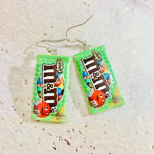 Puede incluir: Un par de pendientes de barra de chocolate M&M's de color verde y amarillo con ganchos plateados. Los pendientes están hechos de arcilla polimérica y tienen la forma de una barra de chocolate M&M's en miniatura. La barra de chocolate tiene las palabras "Crispy" y "M&M's" impresas en ella.