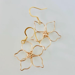 May include: A pair of gold-toned wire earrings, each featuring a five-petaled flower shape constructed from thin, continuous metal wire. The earrings include standard hook-style ear wires for attachment, showcasing a delicate, open-frame design.