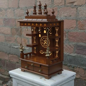 May include: A wooden temple with intricate carvings and a warm, brown finish. The design includes shelves, drawers, and decorative elements like bells and a sun symbol. The temple is approximately 24 inches tall.