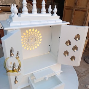 Könnte beinhalten: Ein weißer Holztempel mit offenen Türen, der ein dekoratives Sonnenmuster und das Om-Symbol aufweist. Der Tempel hat eine kleine Schublade und ist mit kleinen Glocken verziert. Die Oberseite hat dekorative Zierelemente.