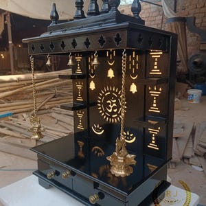 May include: Black wooden temple with intricate cut-out designs, including a sunburst and Om symbol. Features small shelves, hanging bells, and brass accents. The temple has a glossy finish and is approximately 24 inches tall.
