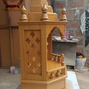 Könnte beinhalten: Holztempel mit aufwendigen Schnitzereien und einem mehrstufigen Design. Die hellbraune Struktur weist dekorative Ausschnitte, gewölbte Eingänge und verzierte Fialen auf. Das Logo "RJ RAJASTHAN ART & CRAFT" ist sichtbar.