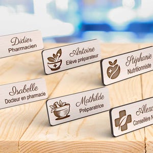 Peut inclure: Plusieurs badges en bois avec des noms et des professions gravés, tels que "Pharmacien" et "Nutritionniste". Chaque badge présente un symbole médical. Les badges sont disposés sur une surface en bois clair.