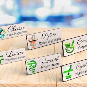 Peut inclure: Plusieurs badges nominatifs en bois avec des bordures noires, affichant des noms et des titres en écriture élégante. Chaque badge présente une icône liée à la pharmacie en vert, bleu sarcelle et rouge, comme un mortier et un pilon ou une croix médicale. Les noms sont Clara, Sylvie, Carine, Lucas, Vincent et Cynthia.
