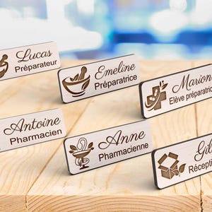Peut inclure: Plusieurs badges en bois gravés avec des symboles de pharmacie et des noms. Chaque badge affiche un nom et un titre différents, comme "Lucas Préparateur" et "Anne Pharmacienne". Les badges sont présentés sur une surface en bois.