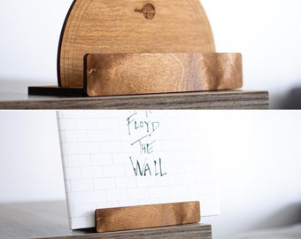 Peut inclure: Un support à disques en bois avec un dessus arrondi et une base rectangulaire. Le support est en bois brun foncé et a une finition naturelle. Le support est conçu pour maintenir un disque vinyle à la verticale. Le support à disques est présenté avec un disque vinyle sur lequel est inscrit le texte "Floyd The Wall".