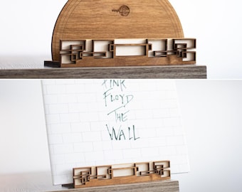 May include: Wooden record stand with a cutout design resembling a brick wall. The stand is holding a white vinyl record with the text "Pink Floyd The Wall" written on it.