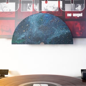 May include: A record player with a vinyl record playing. Above the record player is a piece of art with a galaxy design. Behind the art is a framed album cover with the word "dio" and the words "no angel".