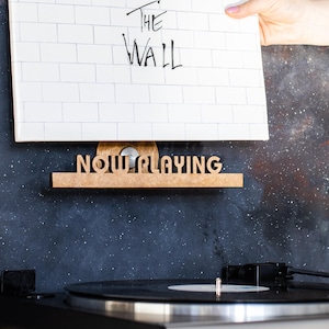 Puede incluir: Un disco de vinilo blanco con el texto "Pink Floyd The Wall" se muestra en un soporte de disco de madera que dice "Now Playing".