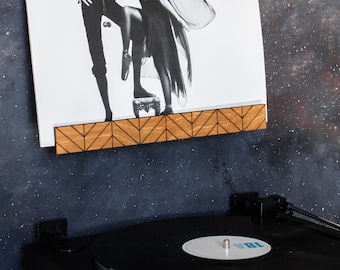May include: A wooden record shelf with a chevron pattern holds a vinyl record with the album cover "Fleetwood Mac Rumours". A black turntable is below the shelf.