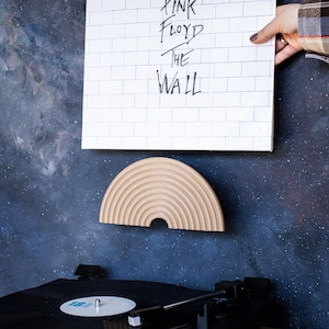 Könnte beinhalten: Ein weißes Albumcover mit dem Text "Pink Floyd The Wall" wird in der Nähe einer halbkreisförmigen Wanddekoration aus Holz gehalten. Ein Plattenspieler mit einer schwarzen Schallplatte befindet sich im Vordergrund. Der Hintergrund ist eine dunkelblaue Wand mit einem Galaxiemuster.