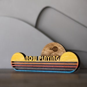 May include: Wooden sign with the words "NOW PLAYING" in a retro font. The sign is decorated with a yellow and orange sunset design and a blue stripe at the bottom.