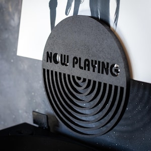 Peut inclure: Un support de tourne-disque circulaire noir avec les mots "NOW PLAYING" découpés. Le support a un design de cercles concentriques.