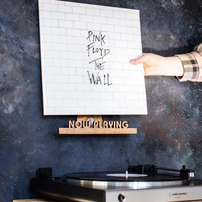 May include: A white vinyl record with the text "Pink Floyd The Wall" on it is displayed on a wooden record holder that says "Now Playing".