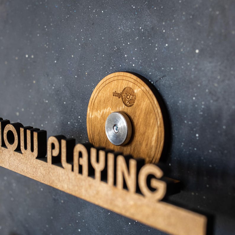 May include: A wooden sign that says "Now Playing" with a round wooden record holder attached to the right side. The record holder has a metal screw in the center and the text "vinyl stone" printed on the surface.