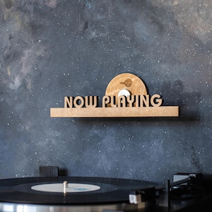 May include: A wooden shelf with the words "NOW PLAYING" in cutout letters. A record player is below the shelf, with a record on the turntable. A white disc with the words "Vinyl Stop" is on the shelf.
