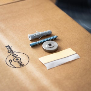 May include: A close-up of a set of hardware for mounting a record player to a wall. The set includes a screw, a wall anchor, a metal mounting plate, and a piece of adhesive tape.
