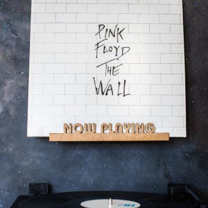 May include: A white vinyl record with the text "Pink Floyd The Wall" written in black marker. A wooden sign that says "Now Playing" is attached to the bottom of the record.