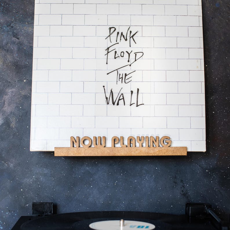 May include: A white vinyl record with the text "Pink Floyd The Wall" written in black marker. A wooden sign that says "Now Playing" is attached to the bottom of the record.