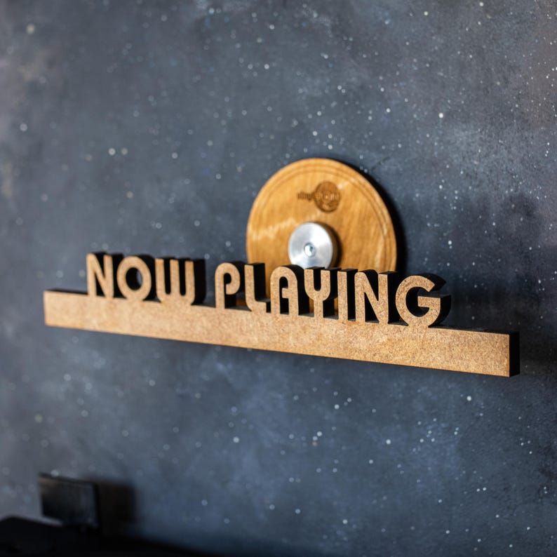 May include: A wooden sign with the words "NOW PLAYING" in black lettering. The sign is mounted on a wall with a wooden disc attached above it.