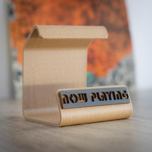 Puede incluir: Un soporte de discos de cartón marrón claro con un recorte negro que dice "now playing".