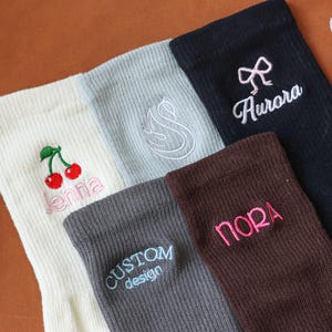 Könnte beinhalten: Fünf Paar Socken mit gestickten Namen und Designs. Die Socken sind weiß, grau, braun und schwarz. Die Designs beinhalten Kirschen, einen Wirbel, eine Schleife und die Worte "Custom Design" und "Nora".