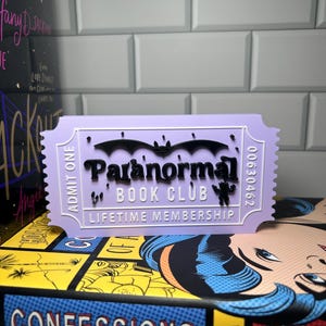 May include: A lavender-colored ticket-shaped item with the words "Paranormal Book Club" and "Lifetime Membership" in black. A bat graphic is above the text. The ticket is on top of a stack of books.