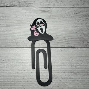 Puede incluir: Un clip negro con un diseño de Ghostface, ojos de corazón rosa y una bebida rosa. El diseño de Ghostface está en la parte superior del clip. El clip está hecho de un material oscuro.