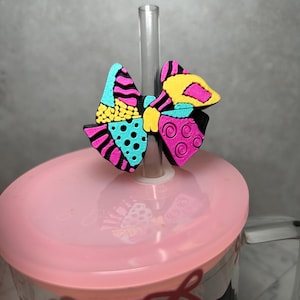 May include: A decorative straw topper shaped like a bow with a vibrant, multi-colored pattern featuring black, bright pink, turquoise, and yellow sections with various textures including dots, swirls, and stripes, attached to a clear plastic straw.