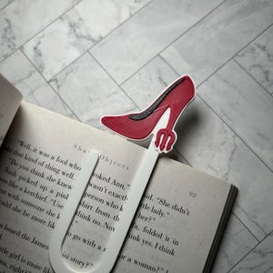 May include: A red high-heeled shoe bookmark with a white base is inserted into a book. The bookmark is made of plastic and has a detailed design. The book is open, with text visible on the pages.
