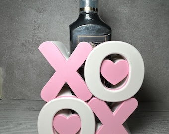 Könnte beinhalten: Ein dekoratives XOXO-Schild in Rosa und Weiß mit Herzakzenten. Hinter dem Schild befindet sich eine schwarze Flasche mit einem Pumpspender. Das Schild steht auf einer hellgrauen Oberfläche.