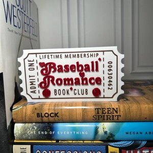 May include: A white ticket-shaped sign with the words "Baseball Romance Book Club" in red, resting on a stack of books. The sign reads "Lifetime Membership" and "Admit One". The books have titles like "Block" and "Teen Spirit".