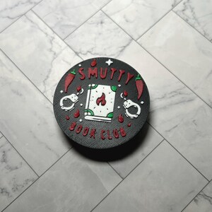 May include: A black round accessory with the text "Smutty Book Club" in red and white. The design includes red chili peppers, handcuffs, and a book with flames. The accessory is likely a phone grip or similar item.