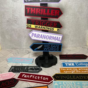 May include: A black vertical post with a base holds multiple arrow-shaped signs in various colours. Signs feature text including Night Court, THRILLER, TRIGGER WARNINGS, PARANORMAL, NO SAFE WORDS, fanfiction, HORROR, WINGS & SHADOWS, and COZY CORNER.