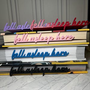 Puede incluir: Una pila de libros con marcapáginas decorativos. Los marcapáginas, en morado, purpurina roja, rosa, azul y negro, dicen "fell asleep here". Los libros tienen páginas amarillas y blancas.