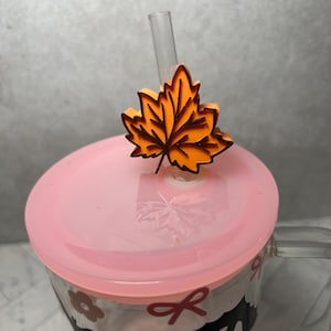 Op de afbeelding: Een doorzichtige beker met een roze deksel en een transparant rietje met een oranje esdoornblad als topper. De beker is versierd met een patroon van zwarte vleermuizen en bruine bloemen en is voorzien van een handvat aan de zijkant van de beker.