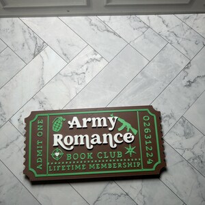 Może przedstawiać: Brązowy i zielony znak w kształcie biletu. Na znaku widnieje napis "Army Romance Book Club Lifetime Membership" białymi literami. Znak ma zieloną obw&oacute;dkę i przedstawia granat i karabin.