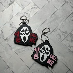 May include: Two ghost-faced character keychains with heart-shaped eyes, each featuring a telephone handset and the text "CALL ME". One keychain has red accents, while the other has light pink accents. Both include a silver-toned metal ball chain.