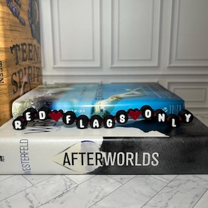 Op de afbeelding: Een zwart kralenarmband met witte letterkralen met de tekst "RED FLAGS ONLY" en rode hartkralen. De armband ligt op een boek met de titel "AFTERWORLDS".