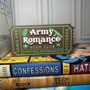 Może przedstawiać: Brązowo-zielony znak w kształcie biletu z napisem "Army Romance Book Club Lifetime Membership". Znak znajduje się na stosie książek, w tym "Confessions" i "Teen Spirit".