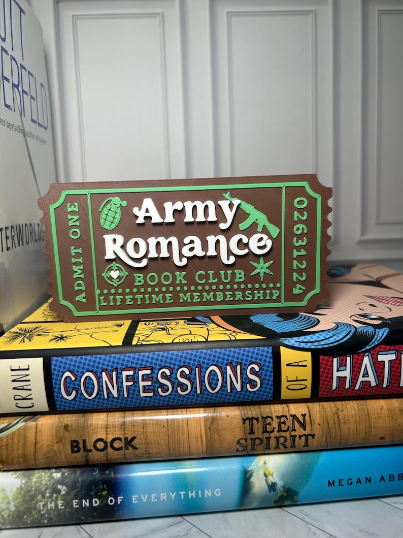 Może przedstawiać: Brązowo-zielony znak w kształcie biletu z napisem "Army Romance Book Club Lifetime Membership". Znak znajduje się na stosie książek, w tym "Confessions" i "Teen Spirit".